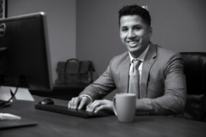 Sam Lensing sitting at a desk smiling with a cup of coffee or tea on the desk.